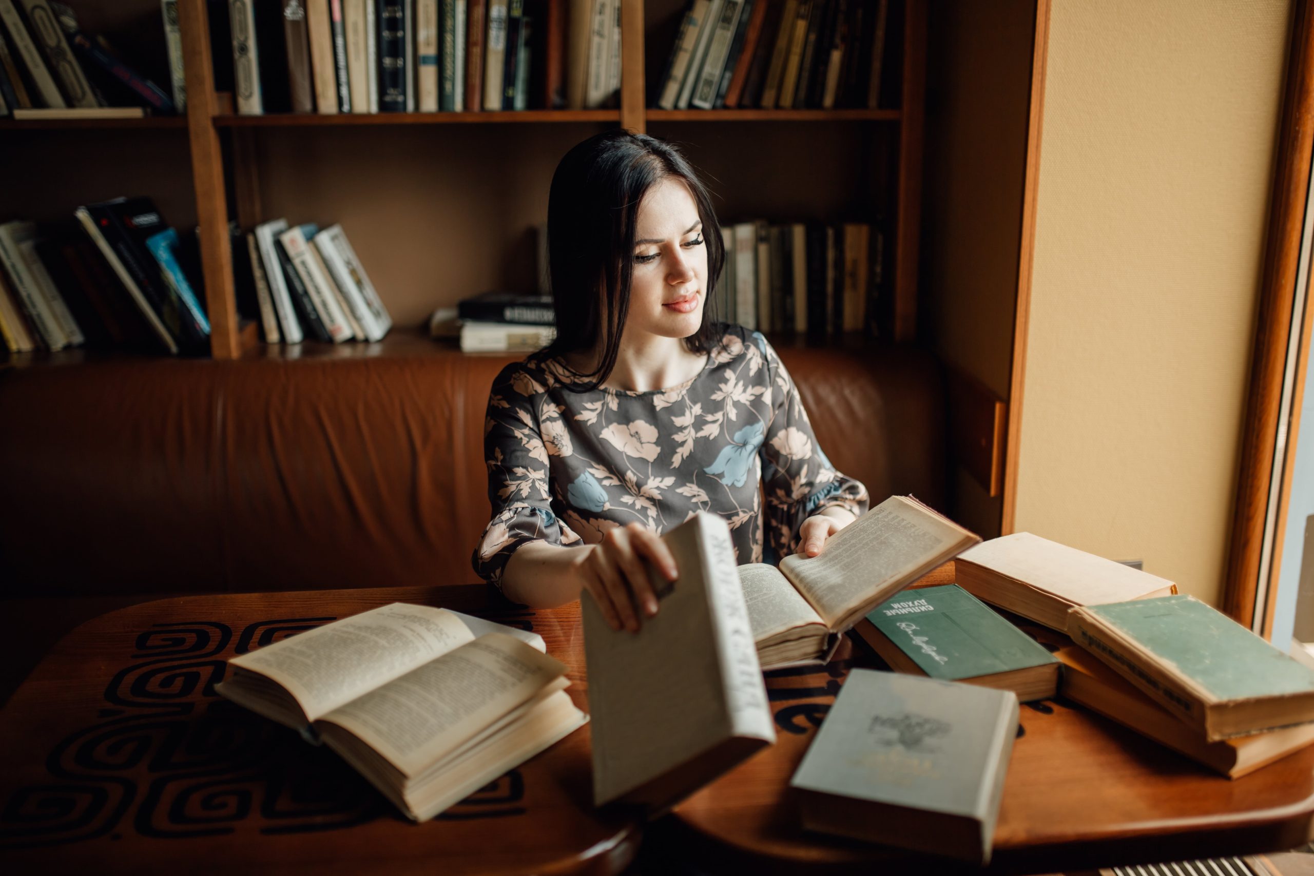 A woman sits at a table surrounded by books, engaged in reading or studying most popular book genres with a focused expression.